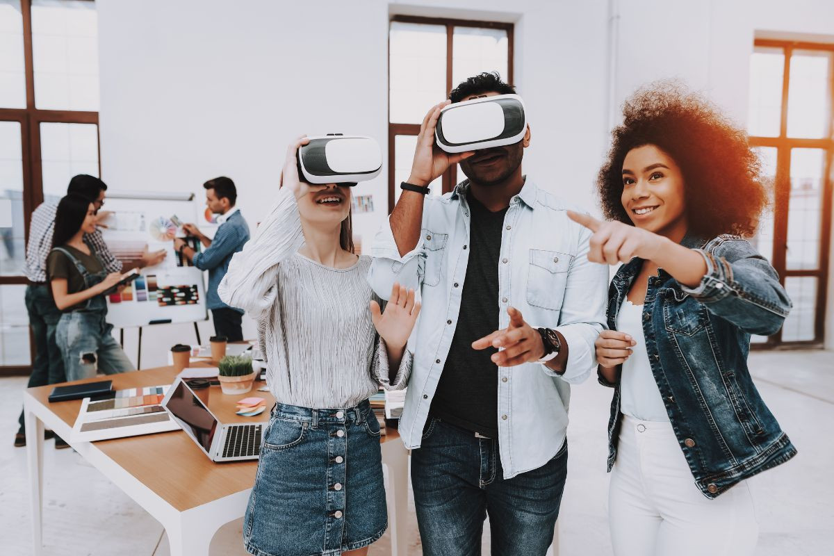 The image shows three people in a modern office space wearing virtual reality (VR) headsets, with one person pointing excitedly. In the background, a few more people are working around a table with laptops and color samples, suggesting a creative or collaborative environment.